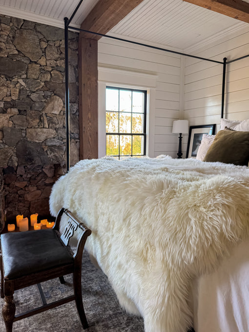 A cozy bedroom features a stone accent wall, window, and a bed topped with the East Perry Sacred Spaces: Plush Ivory Sexto pelt (6’ x 5’). A wooden bench stands at the foot of the bed, with LED candles adding warmth and inviting ambiance.