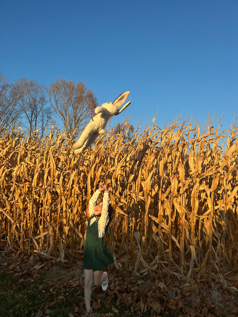 A child in a green dress tosses the East Perry Clover: Shearling Sheepskin Bunny Stuffed with Wool into the air before a dry cornfield, autumn leaves on the ground, under a clear blue sky with bare trees in the background.