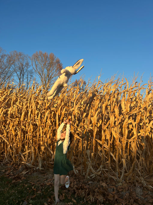 A child in a green dress tosses the East Perry Clover: Shearling Sheepskin Bunny Stuffed with Wool into the air before a dry cornfield, autumn leaves on the ground, under a clear blue sky with bare trees in the background.