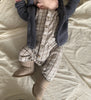 A baby lies on a cream-colored blanket, wearing a plaid jumpsuit, knitted gray cardigan, and East Perry Sheepskin Baby Booties. The baby's face is partially out of frame.