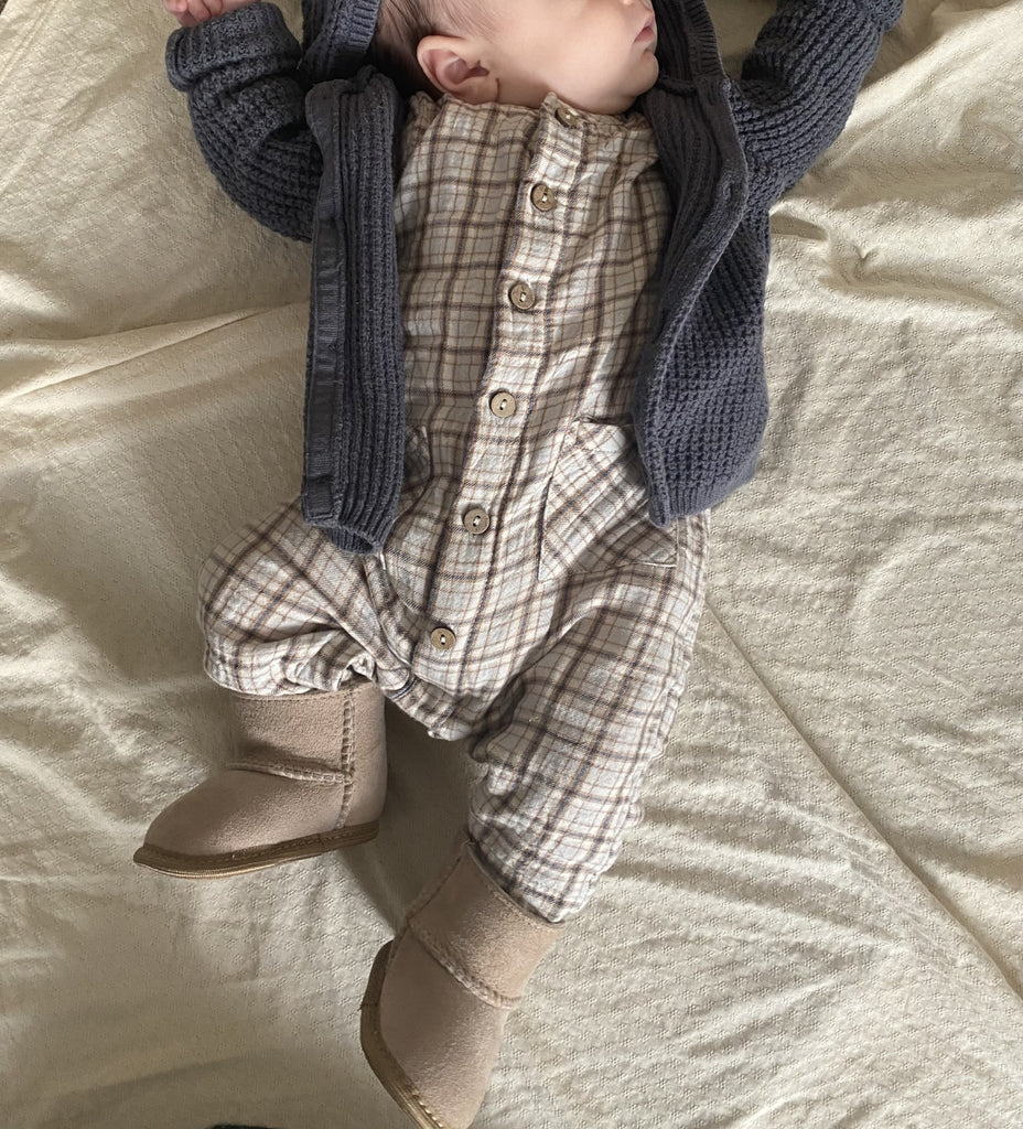 A baby lies on a cream-colored blanket, wearing a plaid jumpsuit, knitted gray cardigan, and East Perry Sheepskin Baby Booties. The baby's face is partially out of frame.