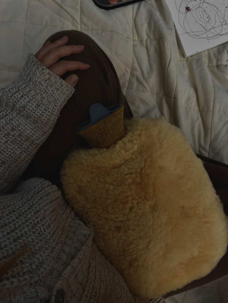 Someone sits on a bed with a white quilted blanket, resting the East Perry Sheepskin Hot Water Bottle Warmer on their lap while wearing a knitted sweater; a sketchbook with drawings lies nearby.