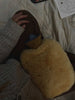 Someone sits on a bed with a white quilted blanket, resting the East Perry Sheepskin Hot Water Bottle Warmer on their lap while wearing a knitted sweater; a sketchbook with drawings lies nearby.