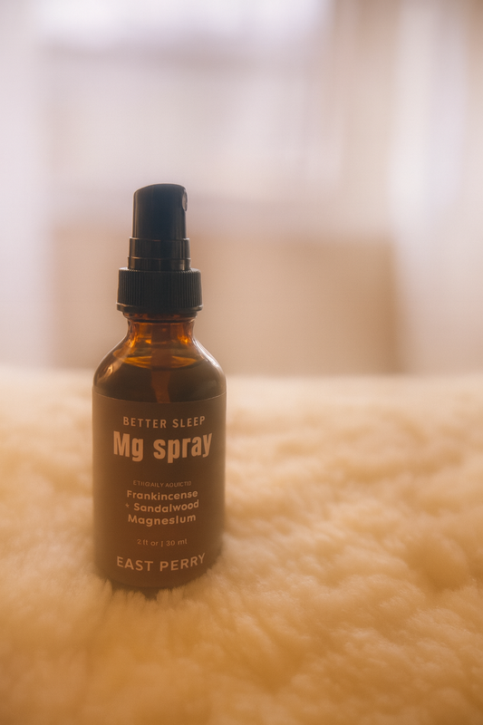 A brown glass spray bottle labeled East Perry Better Sleep Magnesium Spray rests on a soft, fluffy surface with a blurred, warm-toned background.