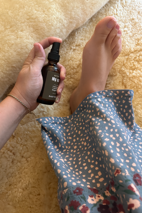 A person holds East Perry Better Sleep Magnesium Spray near their bare foot on a soft cream surface, wearing a blue dress with heart patterns—showcasing the soothing, sleep-supporting benefits of this topical magnesium spray.
