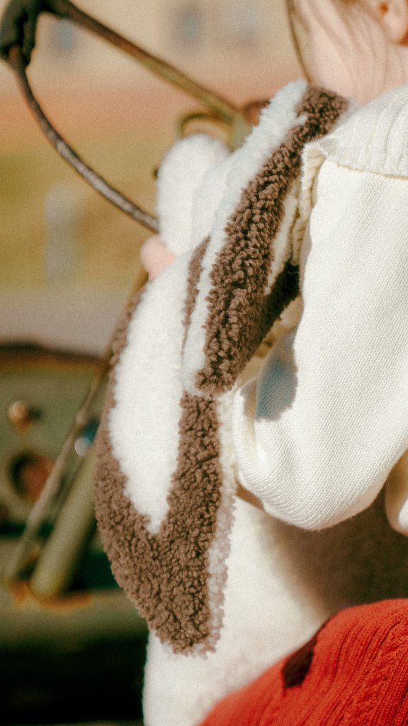 A child in a cozy cream and brown fuzzy jacket holds an old-fashioned steering wheel, with the East Perry Clover Shearling Sheepskin Bunny stuffed with wool peeking from under their arm. The blurred background hints at an outdoor setting.
