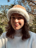 A woman with wavy brown hair smiles outdoors in a white turtleneck and the East Perry Santa Fe Sheepskin Bucket Hat, highlighting cozy winter style against a backdrop of trees and blue sky.