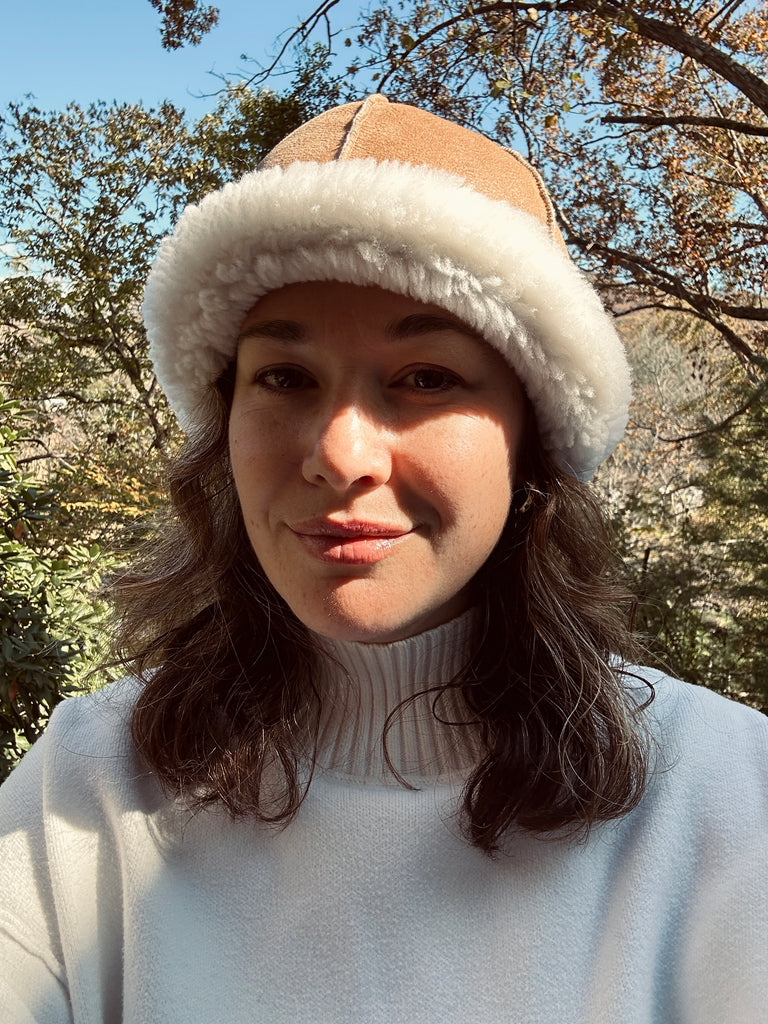 A woman with wavy brown hair smiles outdoors in a white turtleneck and the East Perry Santa Fe Sheepskin Bucket Hat, highlighting cozy winter style against a backdrop of trees and blue sky.