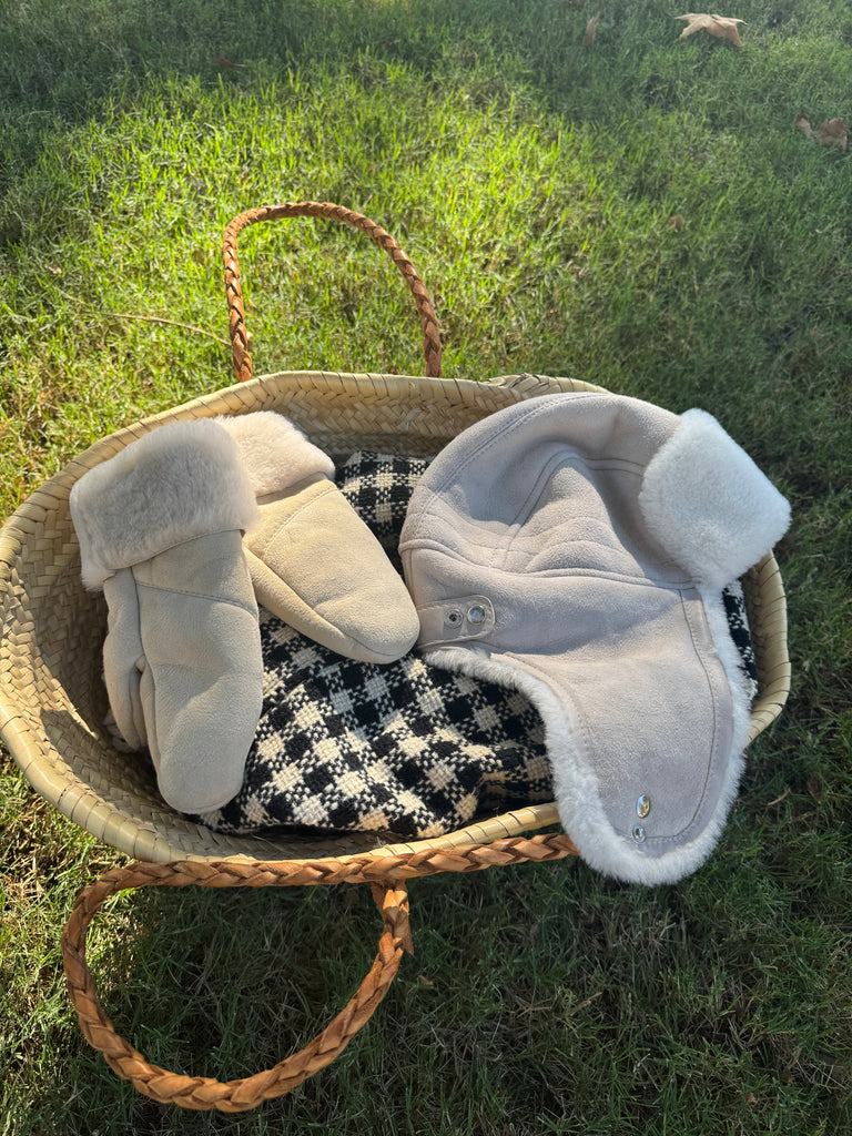 A woven basket on grass holds East Perry’s The Haven Sheepskin Mitten in Cream, warm gloves, a matching shearling-lined hat, and houndstooth fabric. Sunlight casts soft shadows on this cozy scene.