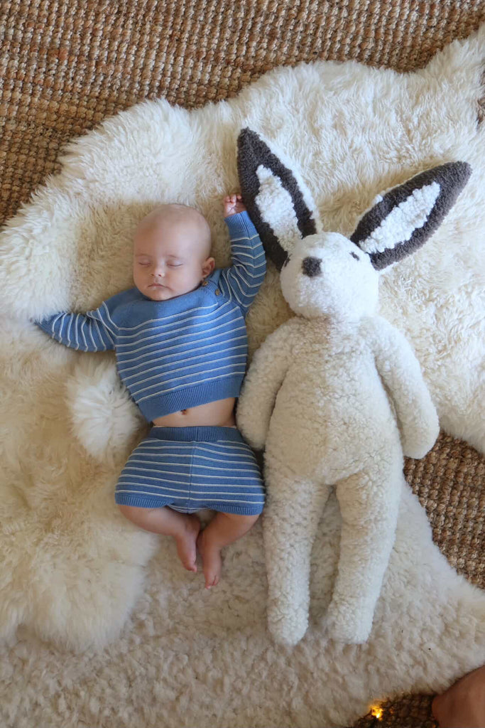 A baby in a blue striped outfit naps on a soft white rug beside the East Perry Clover: Shearling Sheepskin Bunny Stuffed with Wool, both resting on a textured brown carpet.