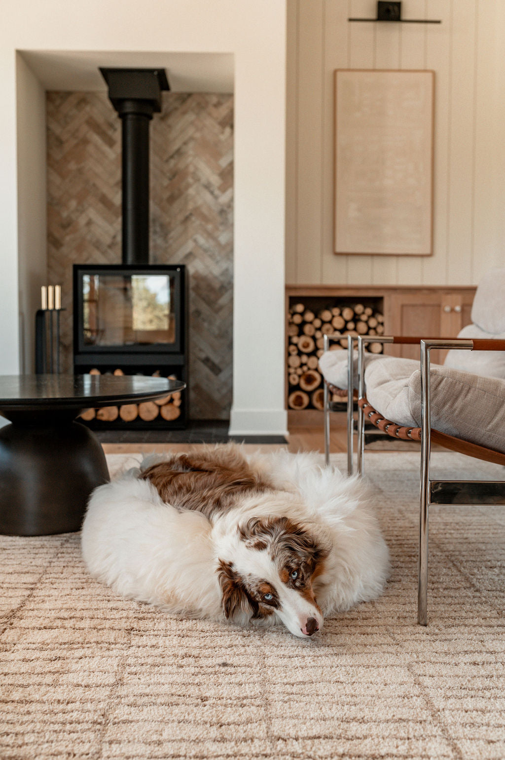 A brown and white dog lies on The Snug in Ivory, Large by East Perryβa fluffy dog bedβin a cozy living room with a modern fireplace, stacked firewood, a round black table, cushioned chairs, and a beige patterned rug.