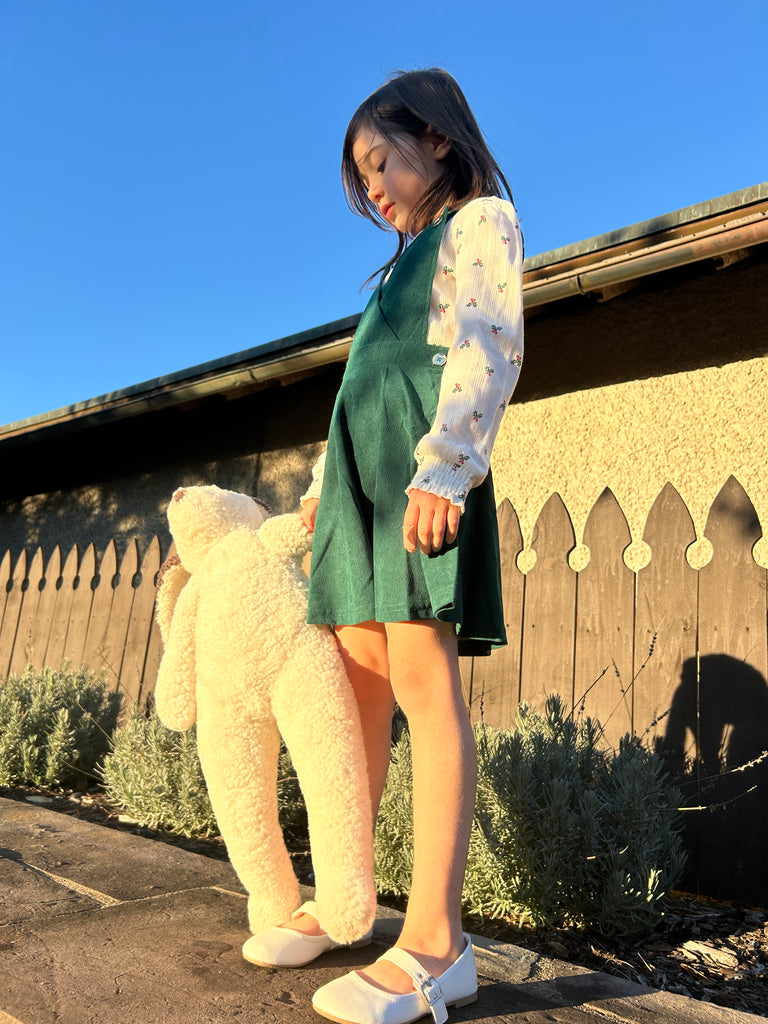 A young girl in a green dress stands outside on a sunny day, holding East Perry’s Clover: Shearling Sheepskin Bunny Stuffed with Wool. She gazes downward, with a wooden fence and shrubs behind her.