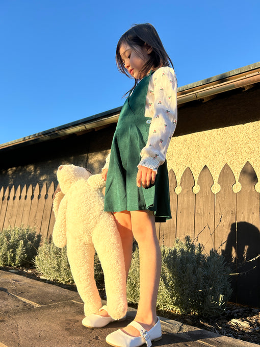 A young girl in a green dress stands outside on a sunny day, holding East Perry’s Clover: Shearling Sheepskin Bunny Stuffed with Wool. She gazes downward, with a wooden fence and shrubs behind her.