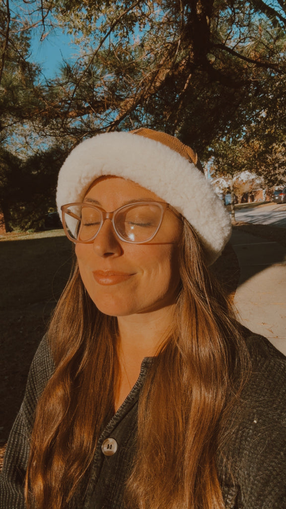 A woman with long brown hair and glasses smiles gently outdoors, wearing the East Perry Santa Fe Sheepskin Bucket Hat—a cozy winter accessory—amid sunlit trees and grass.