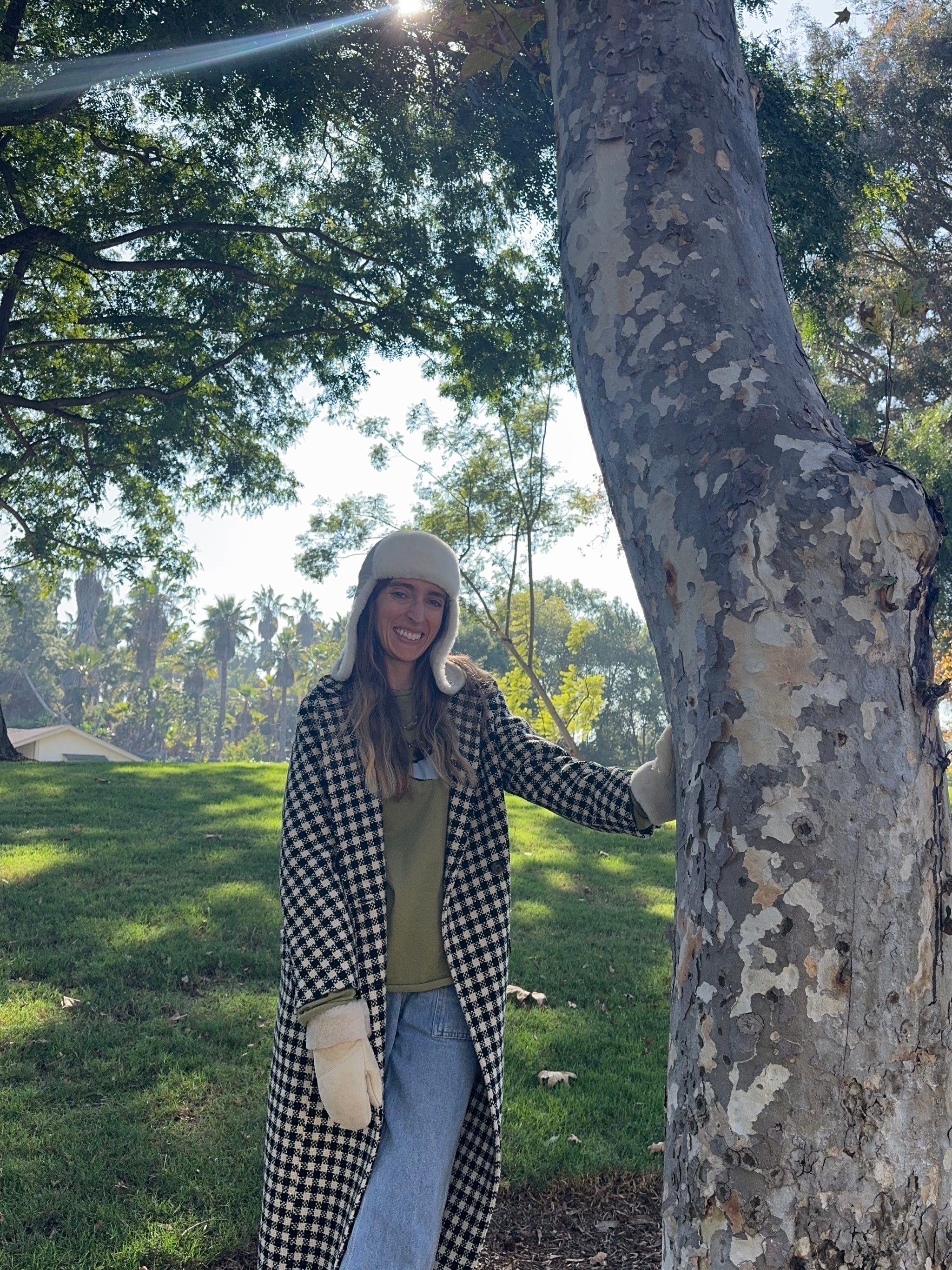 A woman stands outdoors by a tree in a sunlit park, smiling at the camera and wearing East Perry’s The Haven Sheepskin Mitten in Cream, a long checkered coat, and a white beanie. Lush green grass and trees fill the background.