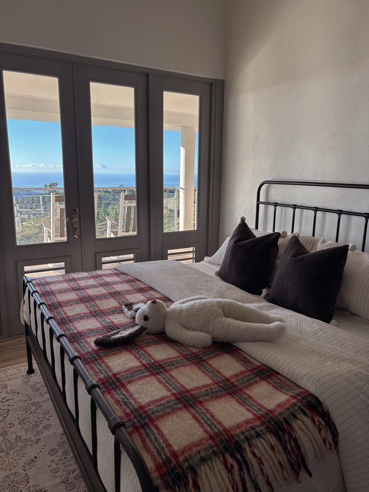 A cozy bedroom features a metal bed frame, plaid blanket, and the Clover: Shearling Sheepskin Bunny Stuffed with Wool by East Perry on the bed. Large French doors lead to a balcony overlooking trees and the ocean beneath a bright blue sky.