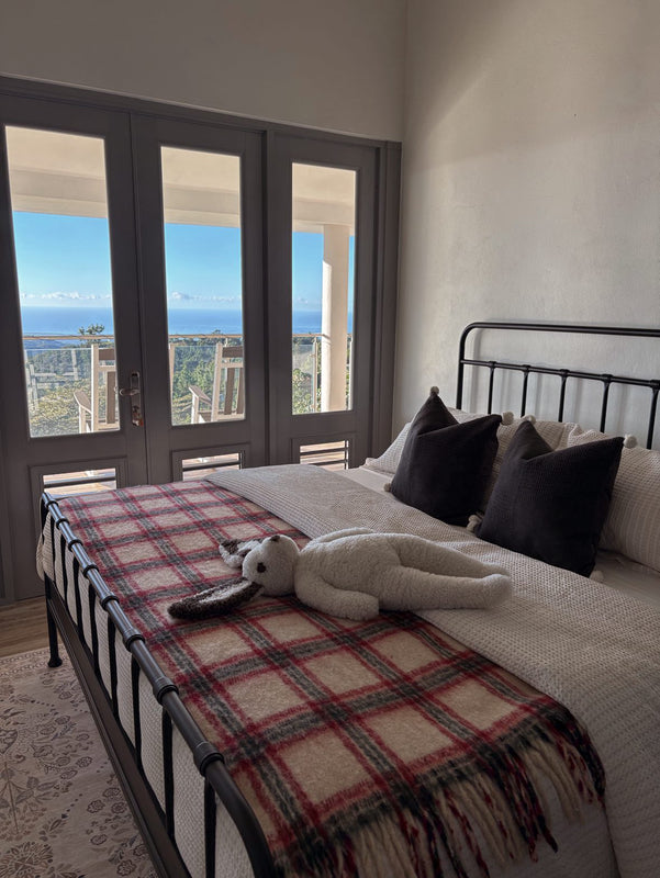 A cozy bedroom features a metal bed frame, plaid blanket, and the Clover: Shearling Sheepskin Bunny Stuffed with Wool by East Perry on the bed. Large French doors lead to a balcony overlooking trees and the ocean beneath a bright blue sky.