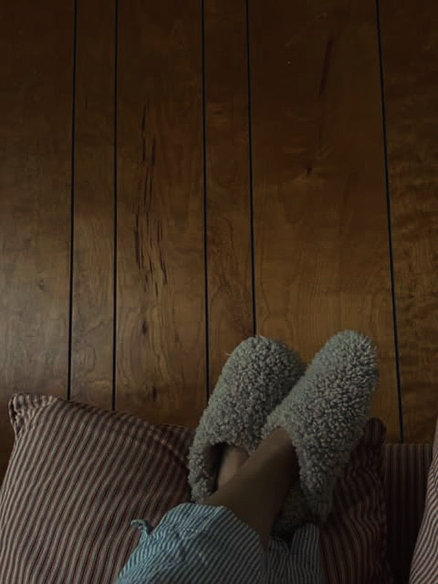 A person wearing East Perry's THE EVERYDAY SLIP-ON: TAN SHEARLING SHEEPSKIN slippers and blue-striped pajama pants rests their feet on a red-and-white-striped pillow against a wooden paneled wall.