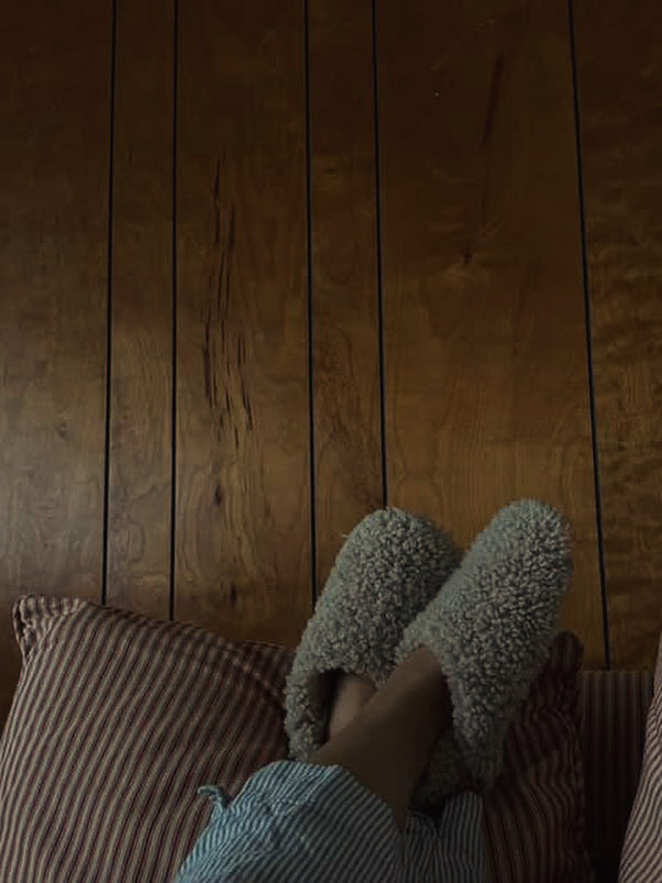 A person wearing East Perry's THE EVERYDAY SLIP-ON: TAN SHEARLING SHEEPSKIN slippers and blue-striped pajama pants rests their feet on a red-and-white-striped pillow against a wooden paneled wall.