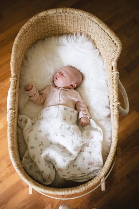 A baby sleeps in a wicker bassinet lined with a soft white blanket and patterned cloth. Beneath, the East Perry Bassinet Sheepskin: Ivory (Small) adds cozy comfort to the wooden floor.