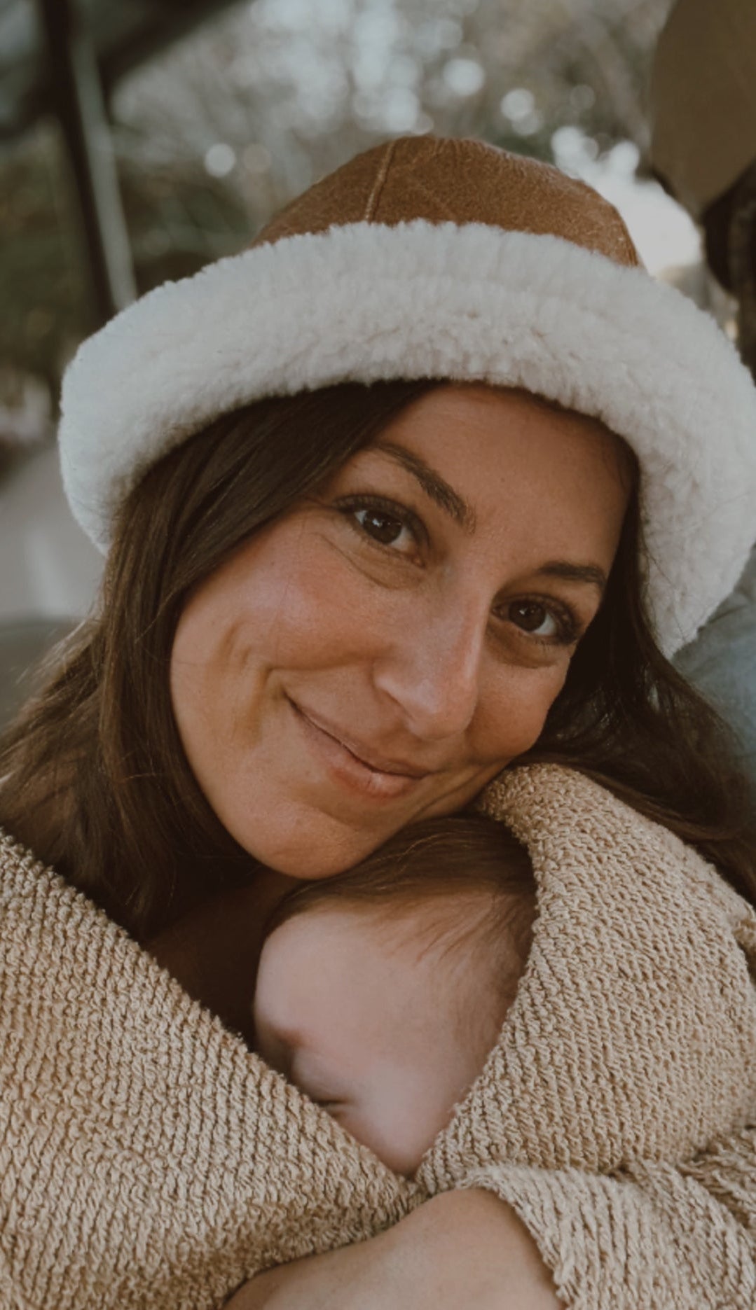 A woman in the East Perry Santa Fe Sheepskin Bucket Hat smiles gently, cradling a baby in a beige blanket close to her chest, evoking winter warmth and style. The baby's face is mostly hidden, adding to the cozy, comforting mood.