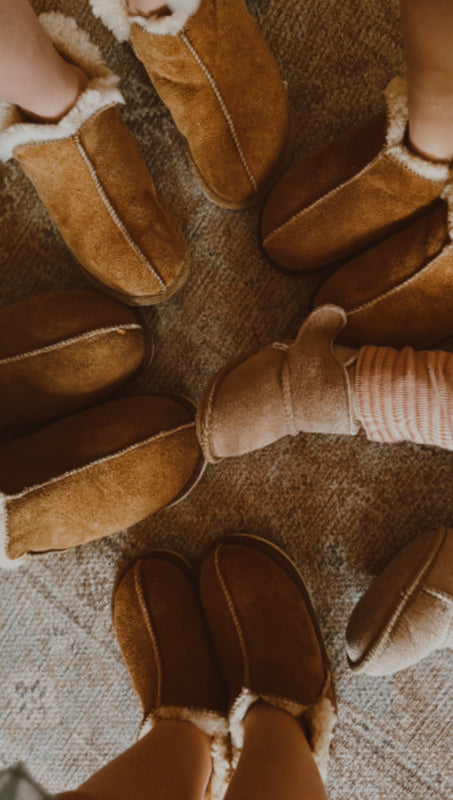 A group stands in a circle on textured carpet, all wearing East Perry Extra Roomy Sheepskin Slipper in Kids' Sizes, their feet pointing toward the center.