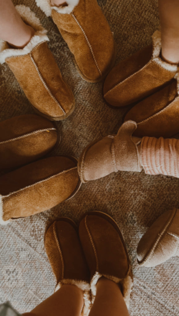 A group stands in a circle on textured carpet, all wearing East Perry Extra Roomy Sheepskin Slipper in Kids' Sizes, their feet pointing toward the center.