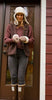 A woman wearing East Perry's The Haven Sheepskin Mitten in Walnut, a furry hat, maroon sweater, brown vest, gray jeans, and rain boots stands before a glass door with a wooden frame against a red exterior wall.