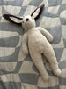 The East Perry Clover: Shearling Sheepskin Bunny Stuffed with Wool features a cream, textured body and long, floppy ears with dark tips, shown lying on a patterned gray and white fabric surface.