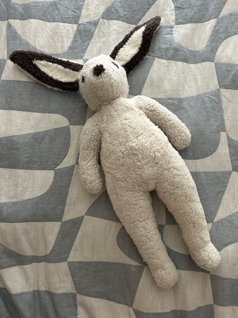 The East Perry Clover: Shearling Sheepskin Bunny Stuffed with Wool features a cream, textured body and long, floppy ears with dark tips, shown lying on a patterned gray and white fabric surface.