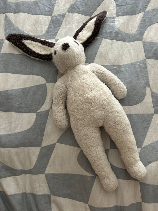 The East Perry Clover: Shearling Sheepskin Bunny Stuffed with Wool features a cream, textured body and long, floppy ears with dark tips, shown lying on a patterned gray and white fabric surface.