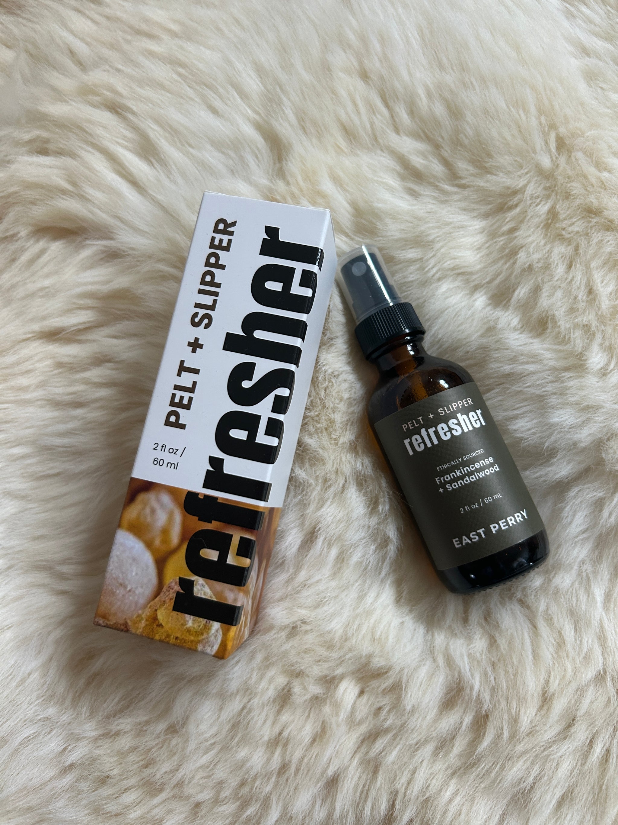 An East Perry Pelt + Slipper Refresher Spray bottle and box sit on soft, cream-colored fur, the bold “refresher” branding evoking its soothing frankincense and sandalwood scent.