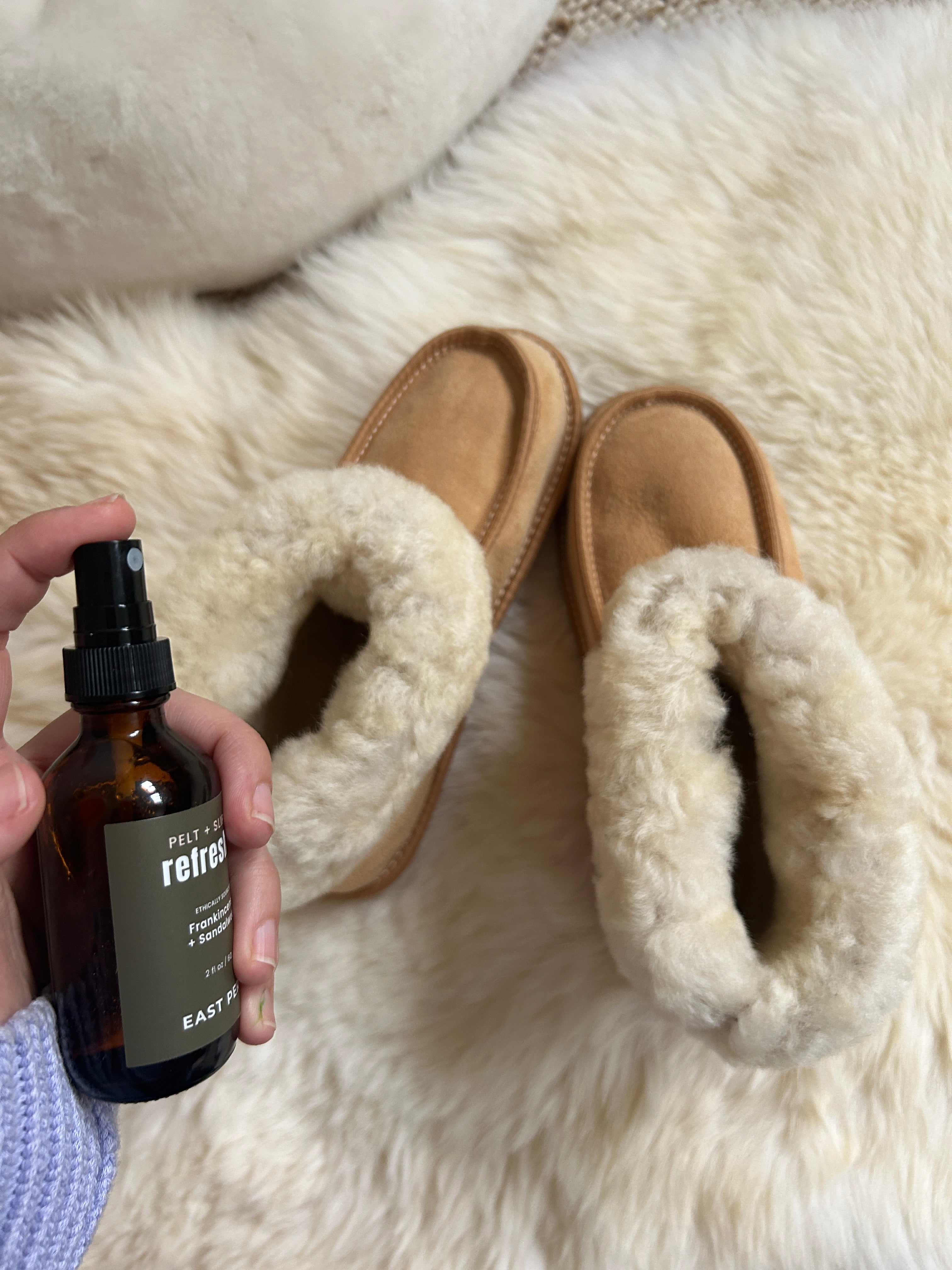 A hand holds East Perry’s Pelt + Slipper Refresher Spray near a pair of tan, fluffy sheepskin slippers on a soft, cream-colored surface.