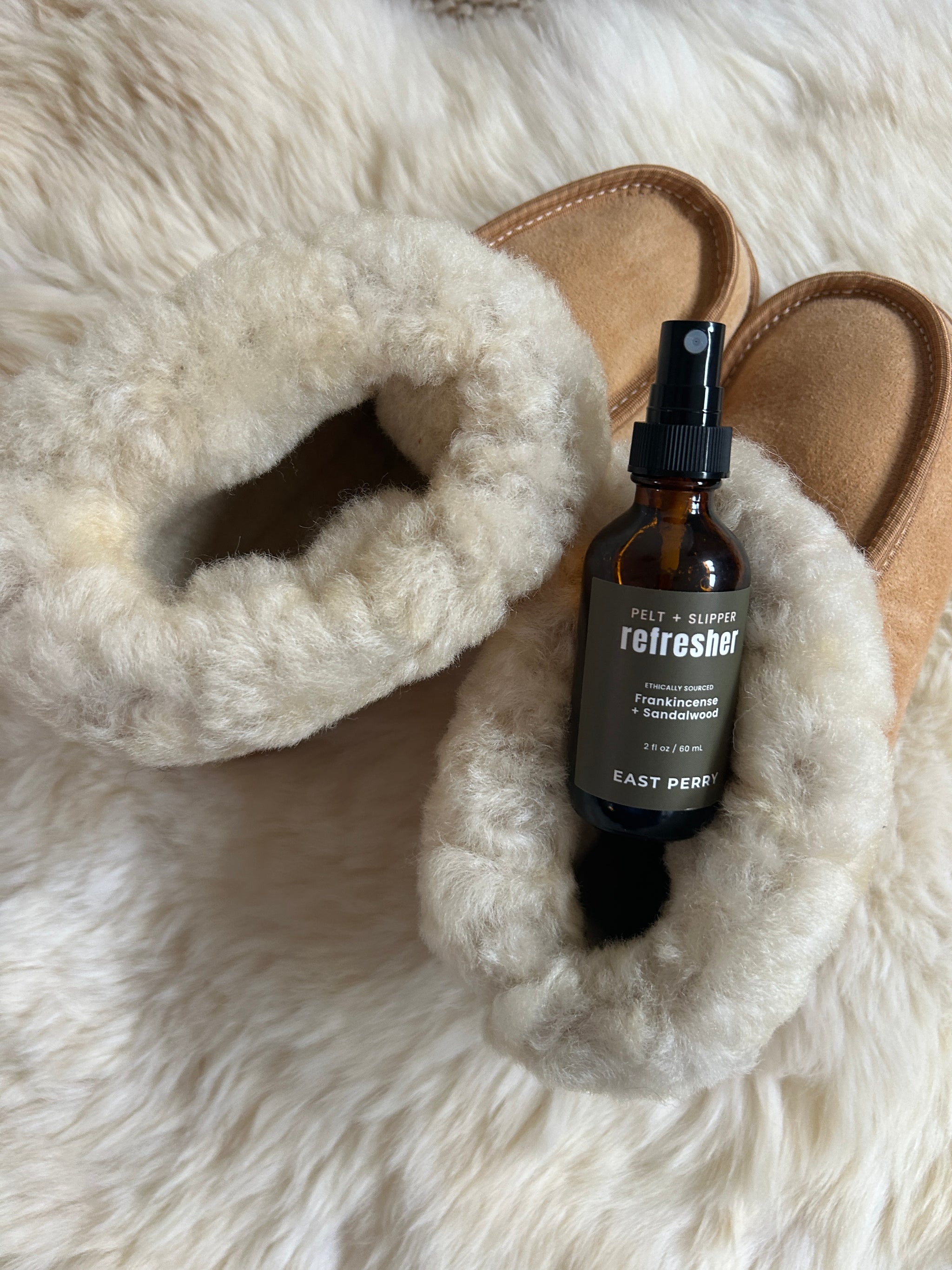 A pair of tan, wool-lined slippers rest on a fluffy white surface with East Perry's Pelt + Slipper Refresher Spray—frankincense sandalwood-scented—placed on top of one slipper.