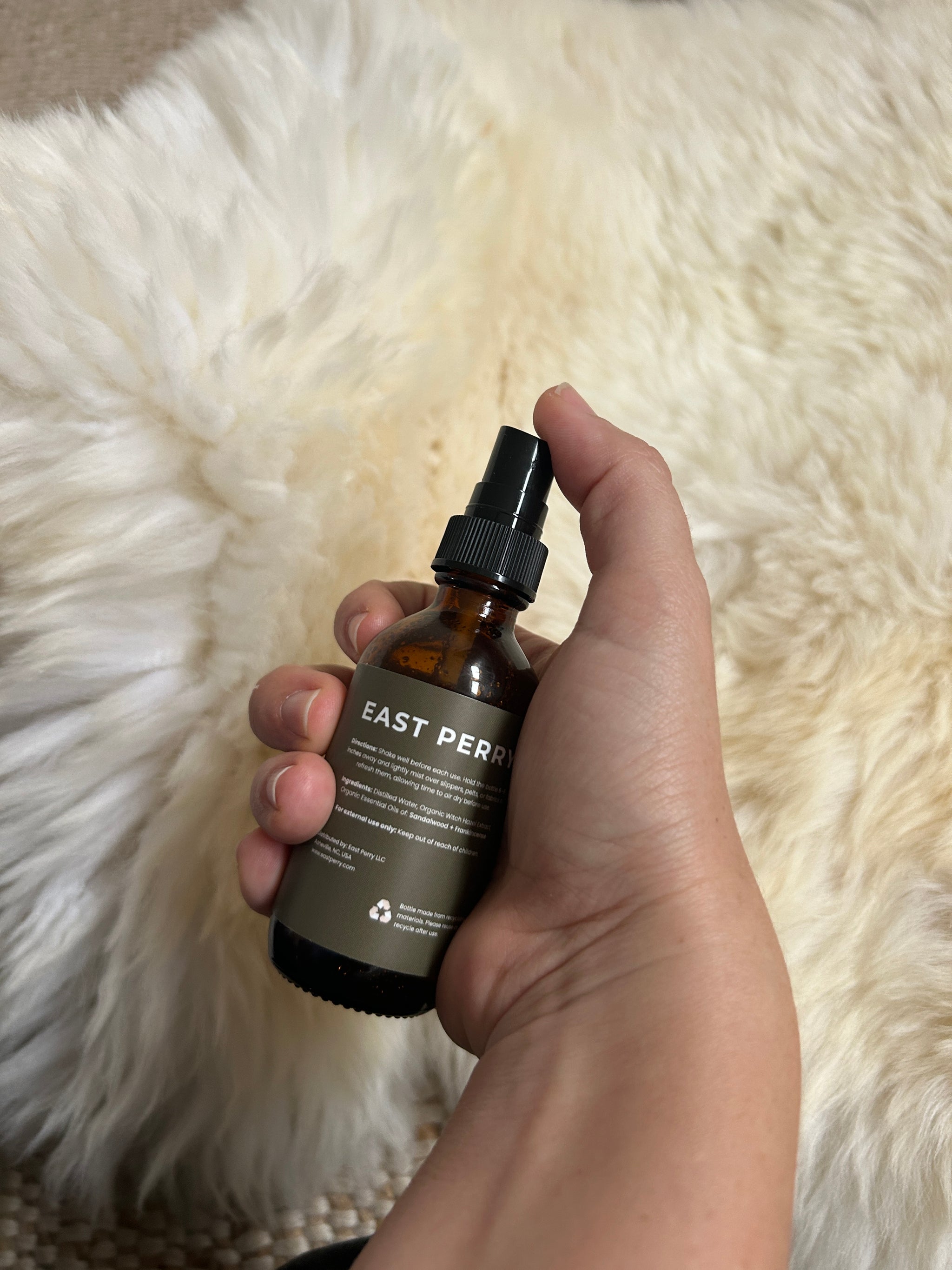 A hand holds a brown glass bottle labeled East Perry Pelt + Slipper Refresher Spray against a soft, cream background—ideal for refreshing sheepskin with notes of frankincense and sandalwood.
