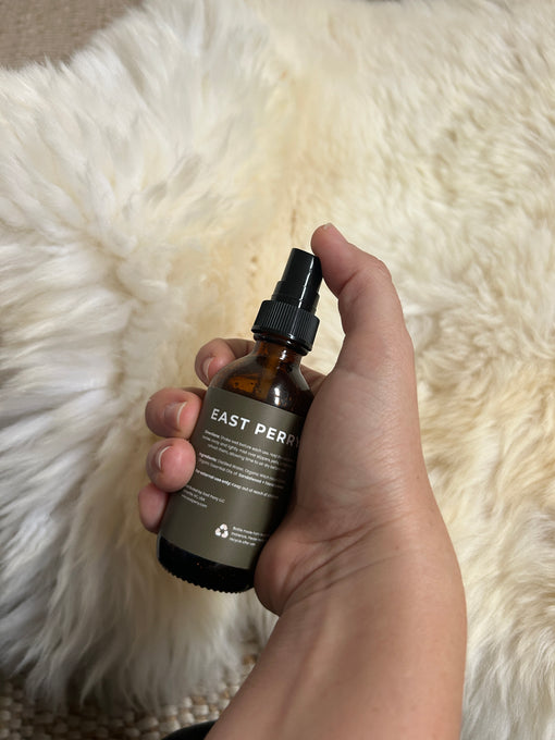 A hand holds a brown glass bottle labeled East Perry Pelt + Slipper Refresher Spray against a soft, cream background—ideal for refreshing sheepskin with notes of frankincense and sandalwood.