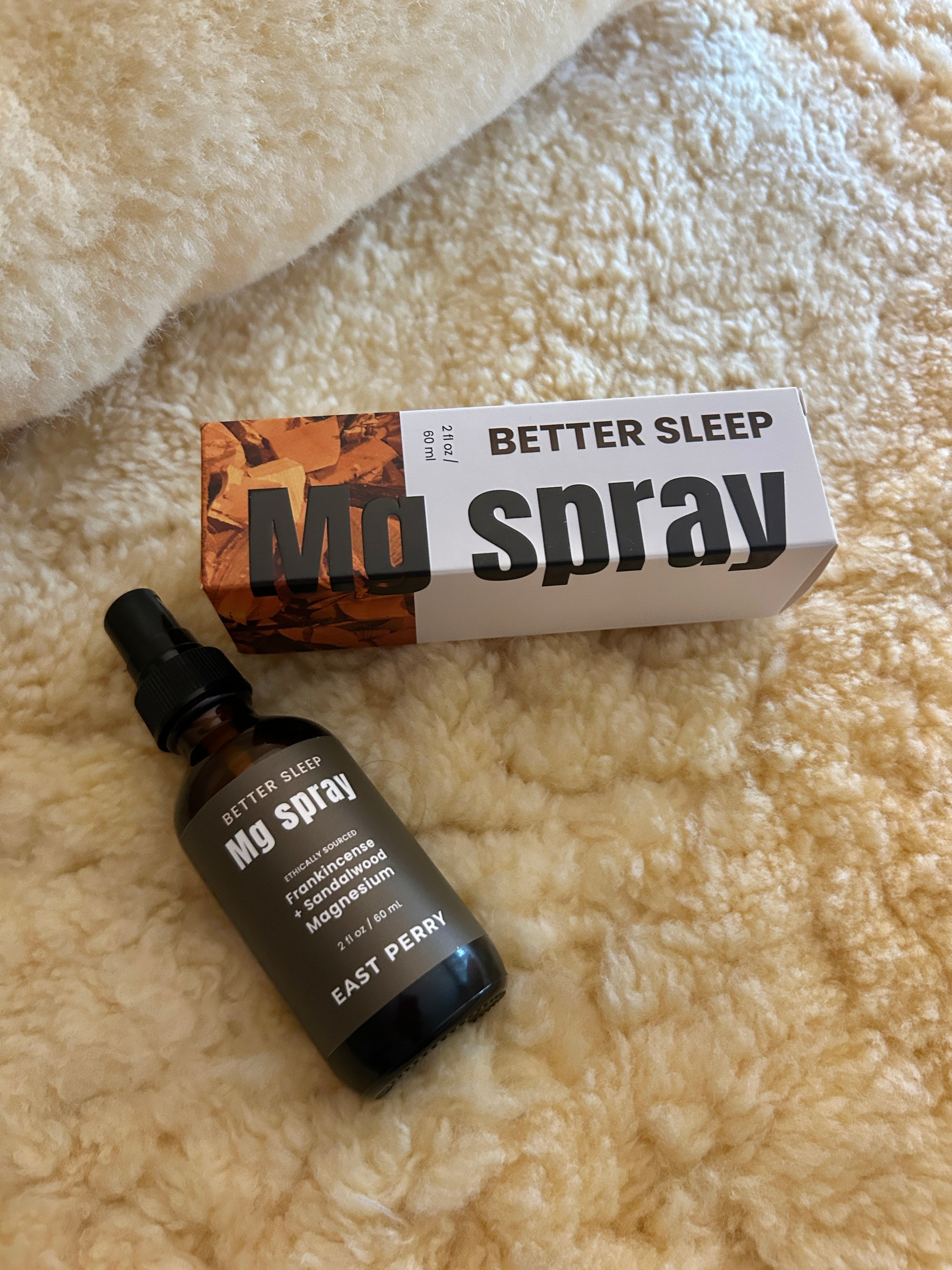 A bottle of East Perry's Better Sleep Magnesium Spray, a topical foot spray for improved sleep, rests on a soft cream surface next to its orange and white box labeled Better Sleep Magnesium Spray.