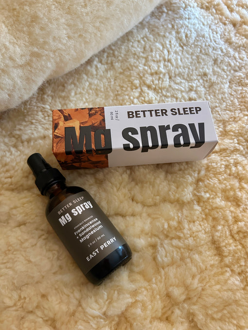 A bottle of East Perry's Better Sleep Magnesium Spray, a topical foot spray for improved sleep, rests on a soft cream surface next to its orange and white box labeled Better Sleep Magnesium Spray.
