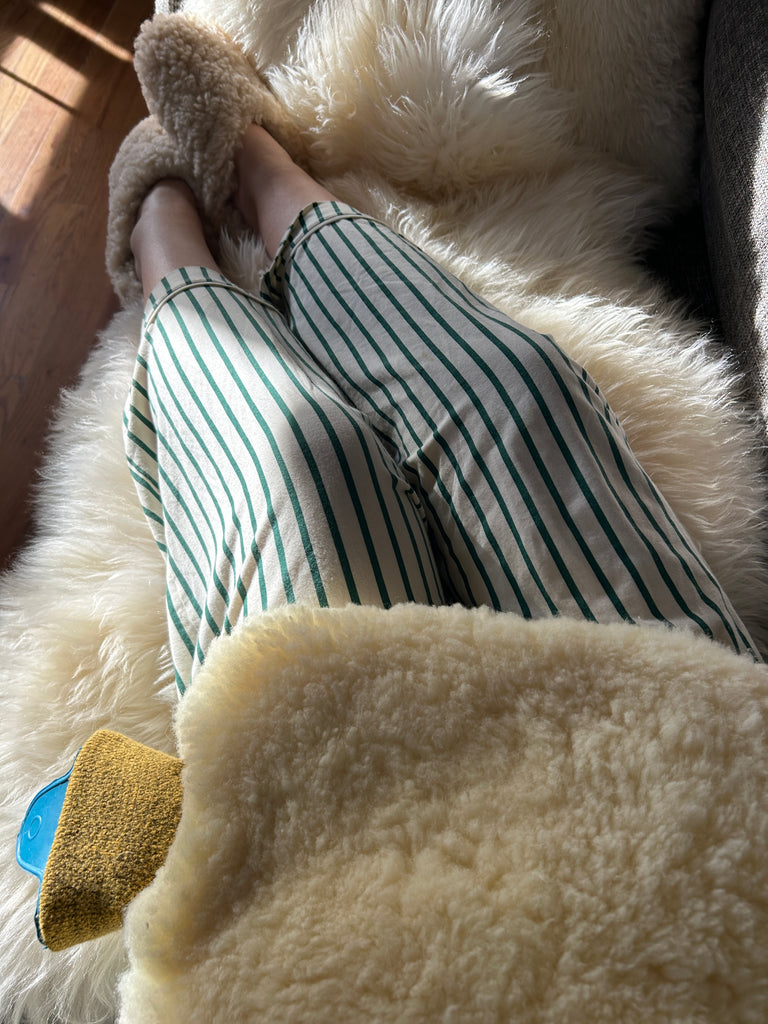 Lounging in sunlight on a plush cream blanket, wearing striped pajama pants and sheepskin slippers, and holding The Reset Bundle by East Perry—everything you need for a cozy nervous system reset.