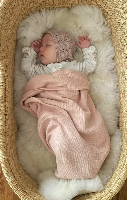 A sleeping baby in a knit bonnet and patterned pajamas rests in a woven basket, nestled on the East Perry Bassinet Sheepskin: Ivory (Small) and covered with a soft pink blanket.