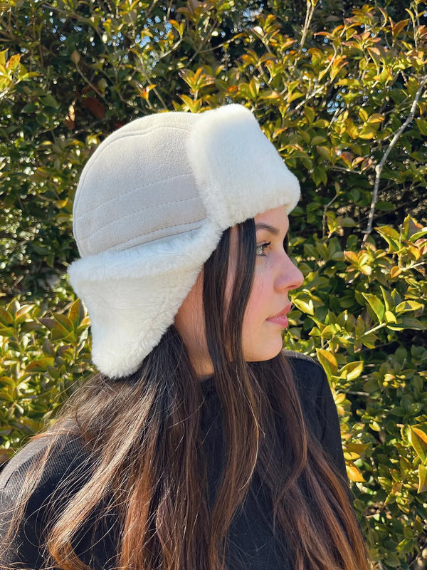 A young woman with long brown hair stands outside by green bushes, wearing the East Perry Sheepskin and Leather Pilot Hat in Cream, featuring white fur trim, as she looks to the right.