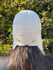 A person with long brown hair, seen from behind, wears the East Perry Sheepskin and Leather Pilot Hat in Cream outdoors in front of leafy green bushes.