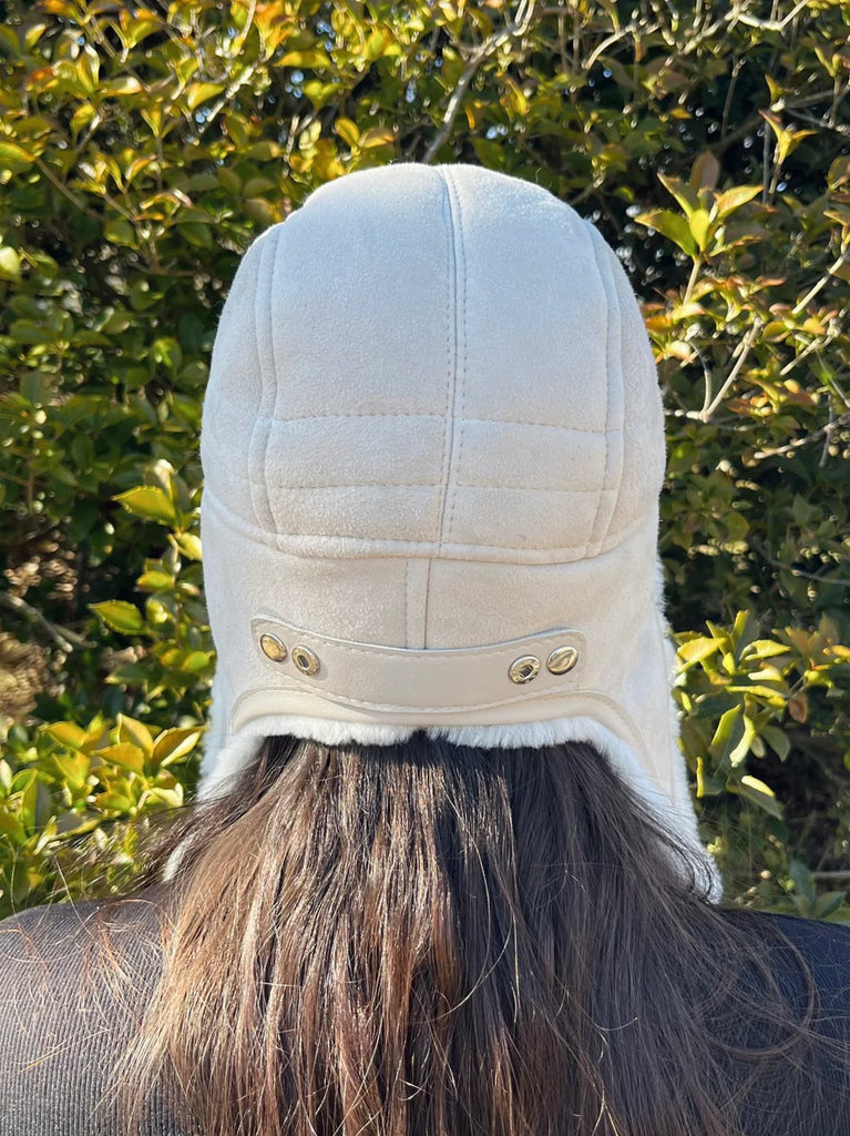 A person with long brown hair, seen from behind, wears the East Perry Sheepskin and Leather Pilot Hat in Cream outdoors in front of leafy green bushes.
