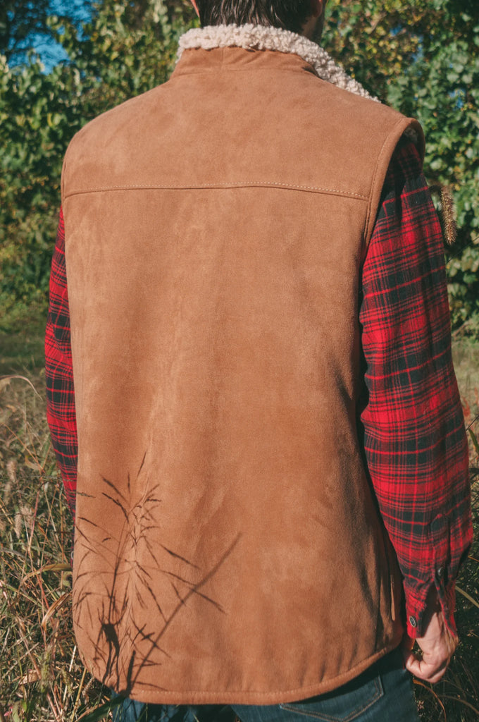 Person wearing a brown sheepskin mens vest over a red plaid shirt in an outdoor setting.